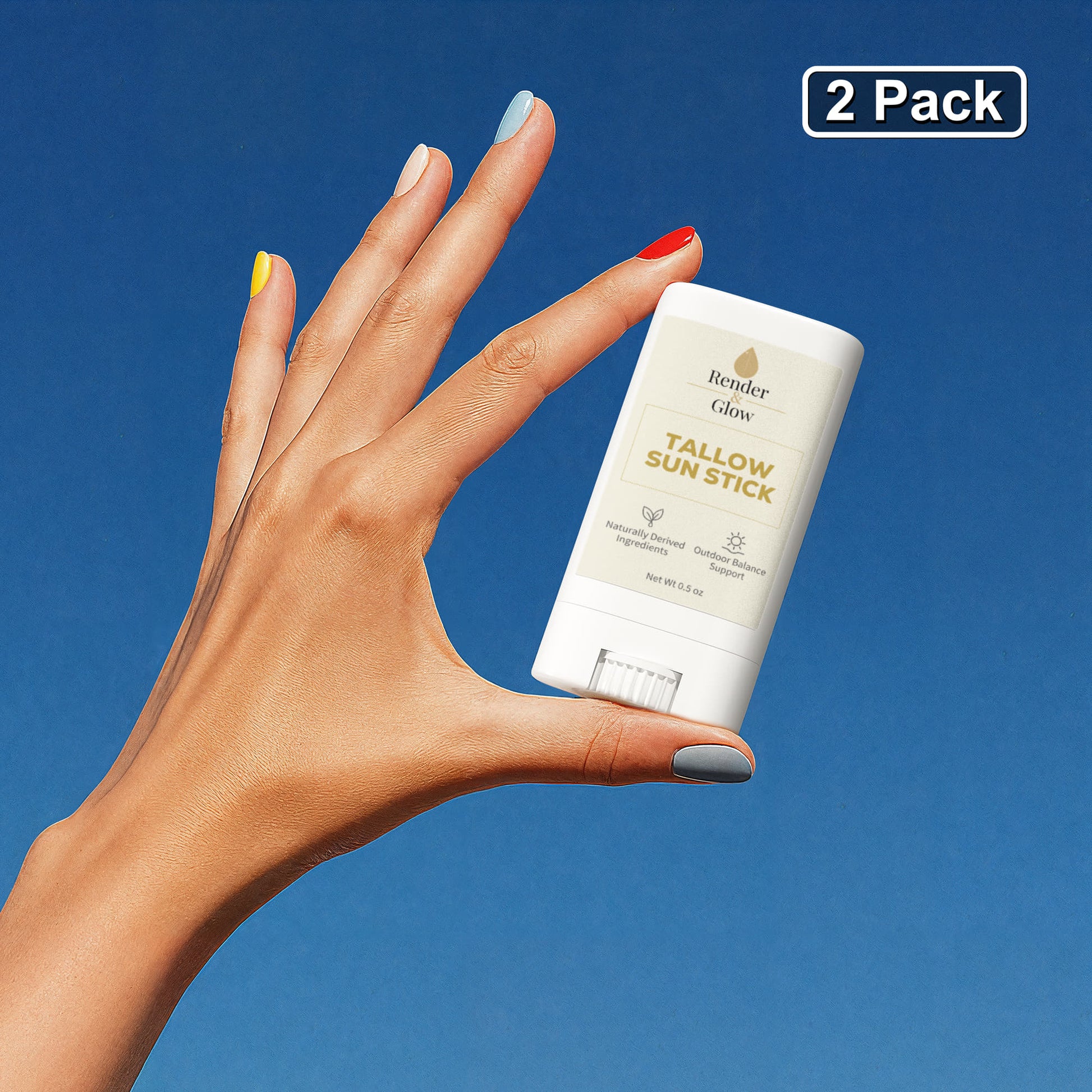 A hand with colorful nails holds a Render & Glow Tallow Sun Stick (2-Pack) with zinc oxide against blue.