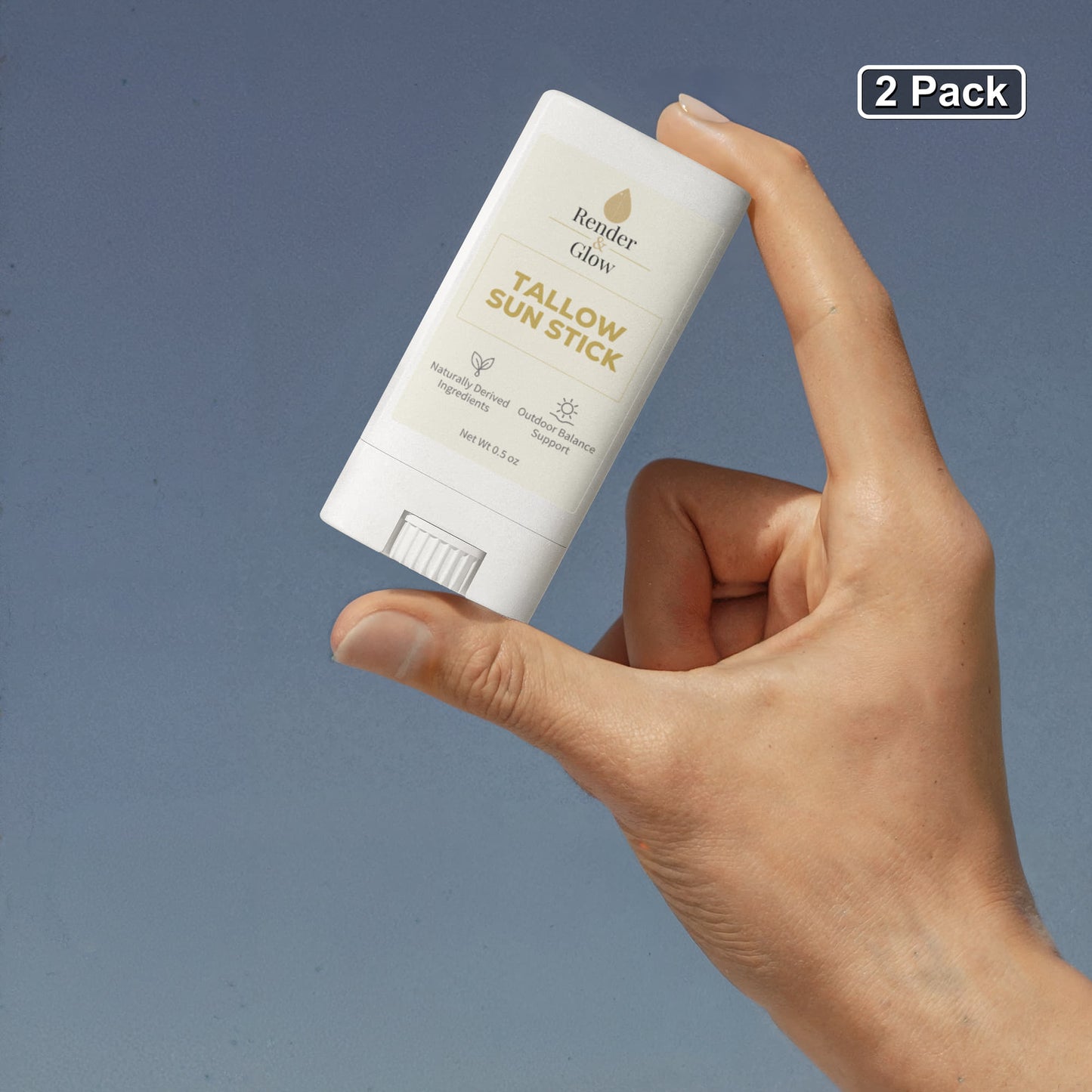A hand holds a Render & Glow Tallow Sun Stick (2-Pack) with zinc oxide against a blue background.