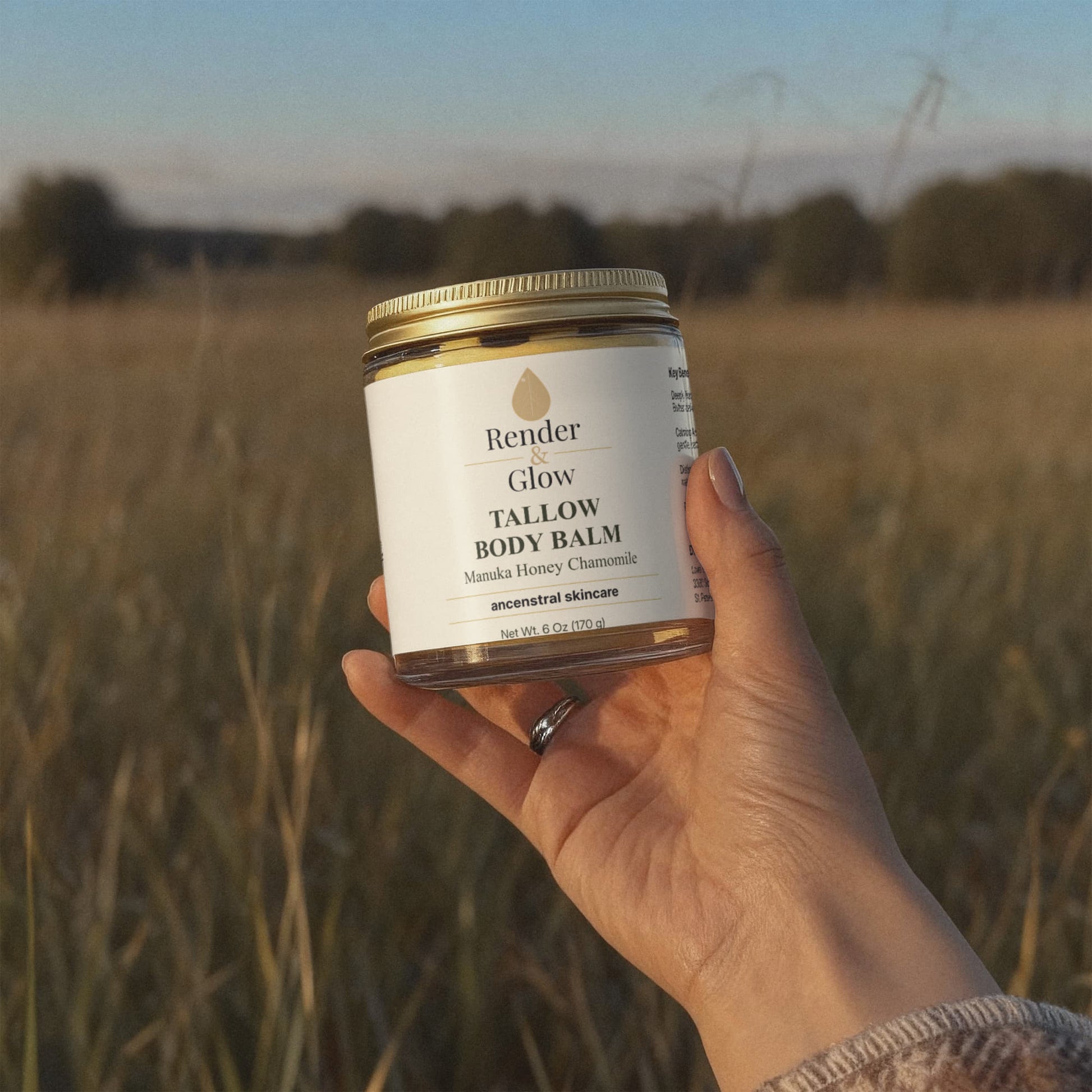 A hand holds a jar of Render & Glow Tallow Body Balm, made with grass-finished beef tallow, in a grassy field at sunset.