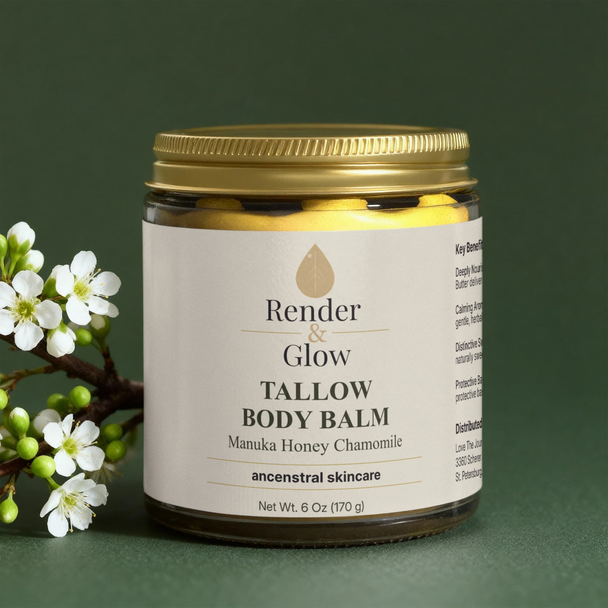 A jar of Render & Glow Tallow Body Balm sits beside white blossoms on a dark green surface.