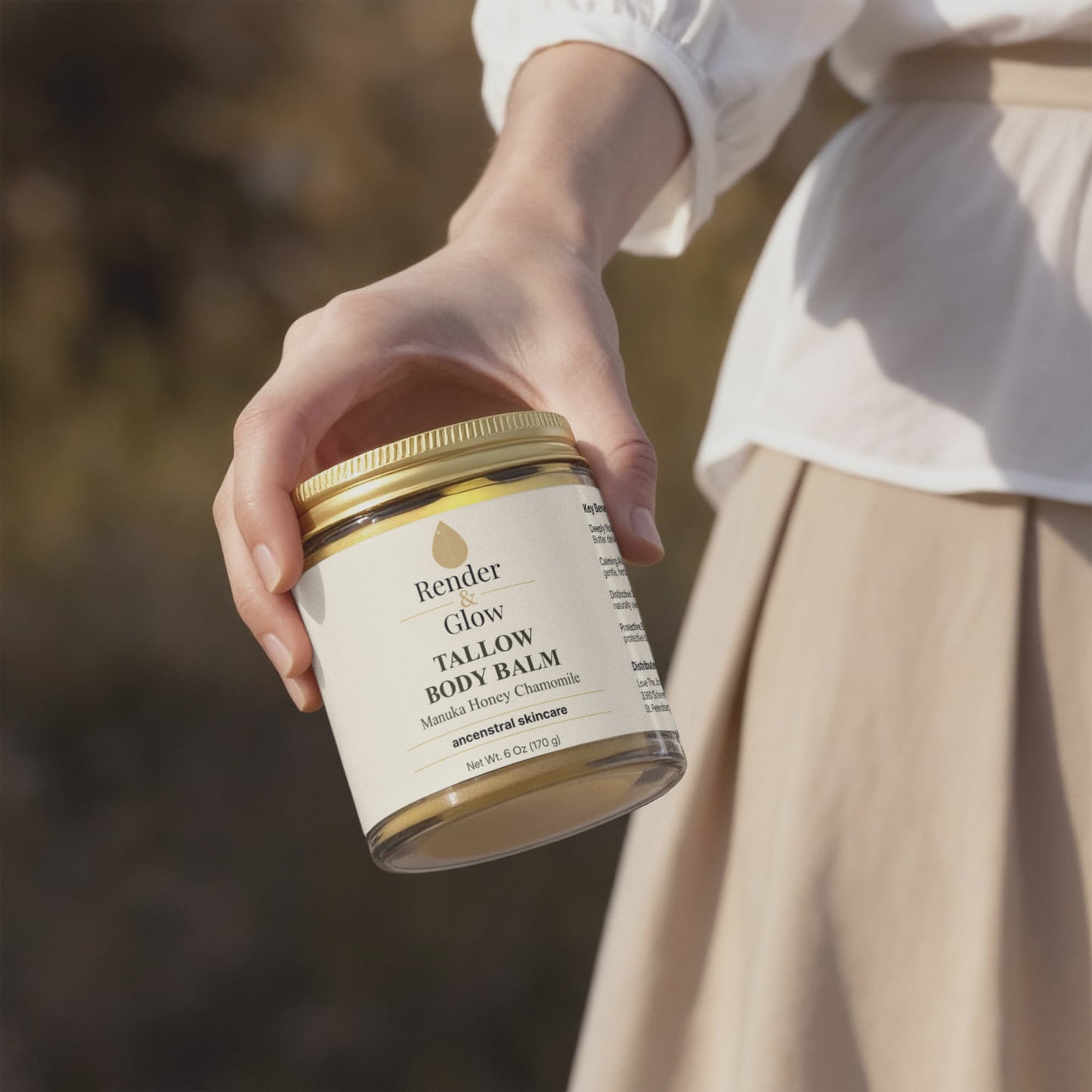 A person holds Render & Glow Tallow Body Balm for intense hydration, outdoors in a white blouse and beige skirt.